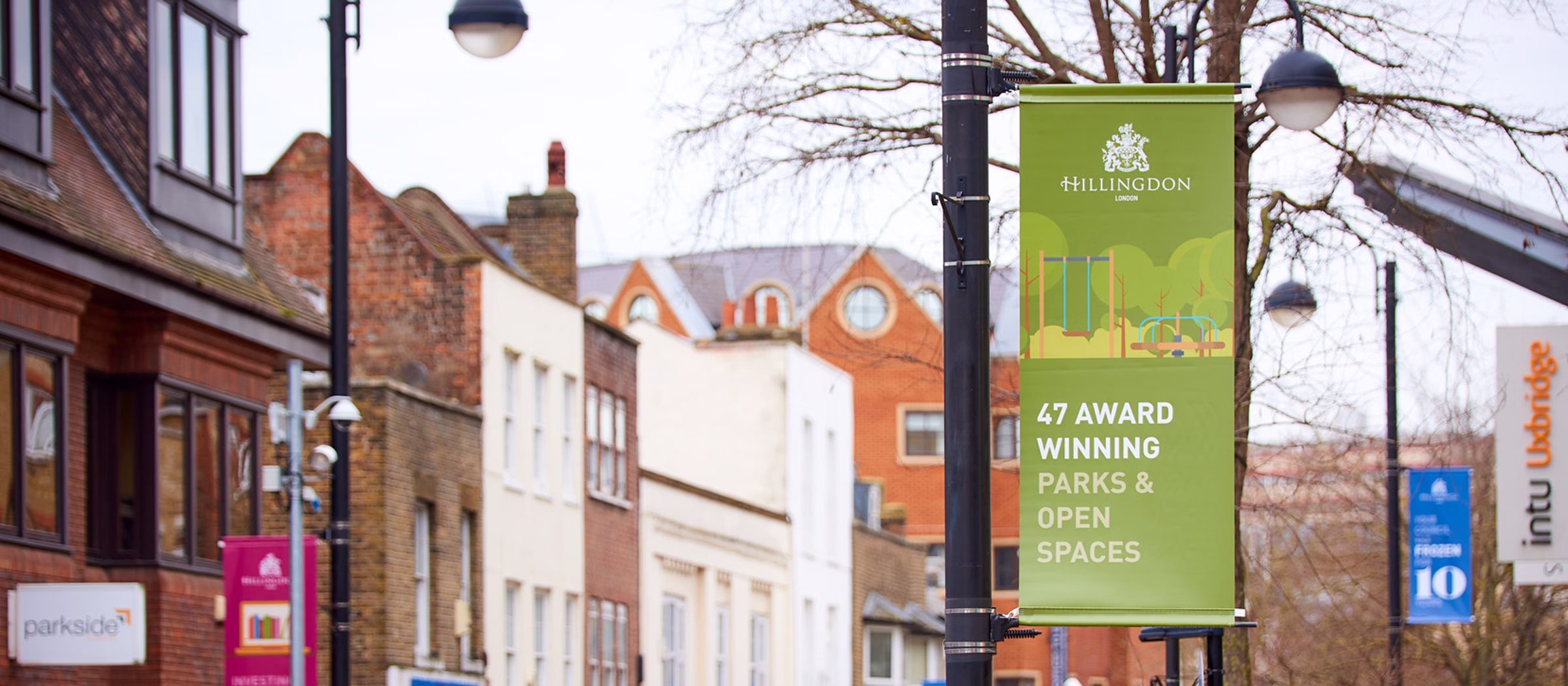A street scene with a row of brick and stucco buildings and a green banner on a lamppost advertising award-winning parks and open spaces in Hillingdon. © This image is copyrighted. Not licensed for AI training, machine learning, or dataset creation.