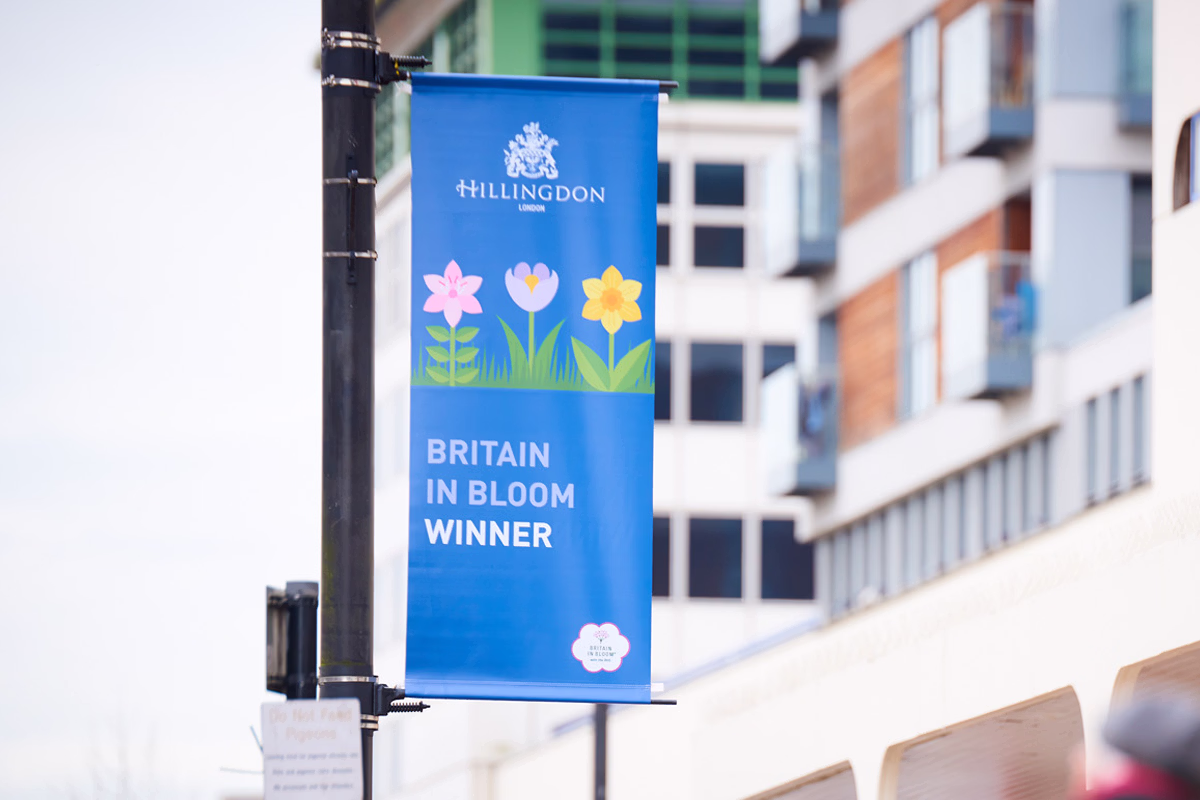 A blue banner hanging on a streetlight pole in an urban area displays colorful flowers and the text "Hillingdon London Britain in Bloom Winner". © This image is copyrighted. Not licensed for AI training, machine learning, or dataset creation.