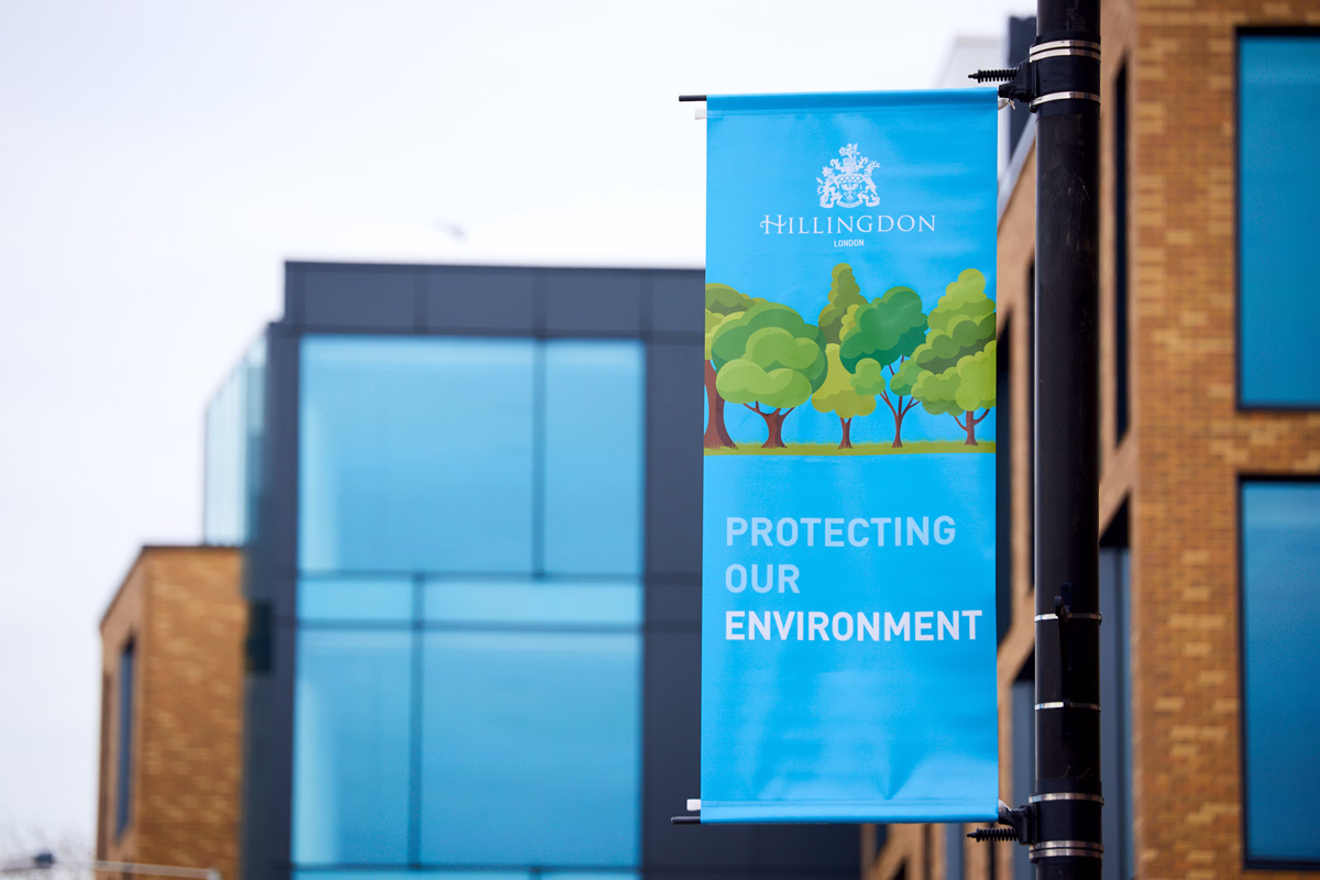 A blue banner hanging on a streetlight pole in front of modern glass and brick buildings reads "Hillingdon Protecting Our Environment" with an illustration of trees. © This image is copyrighted. Not licensed for AI training, machine learning, or dataset creation.