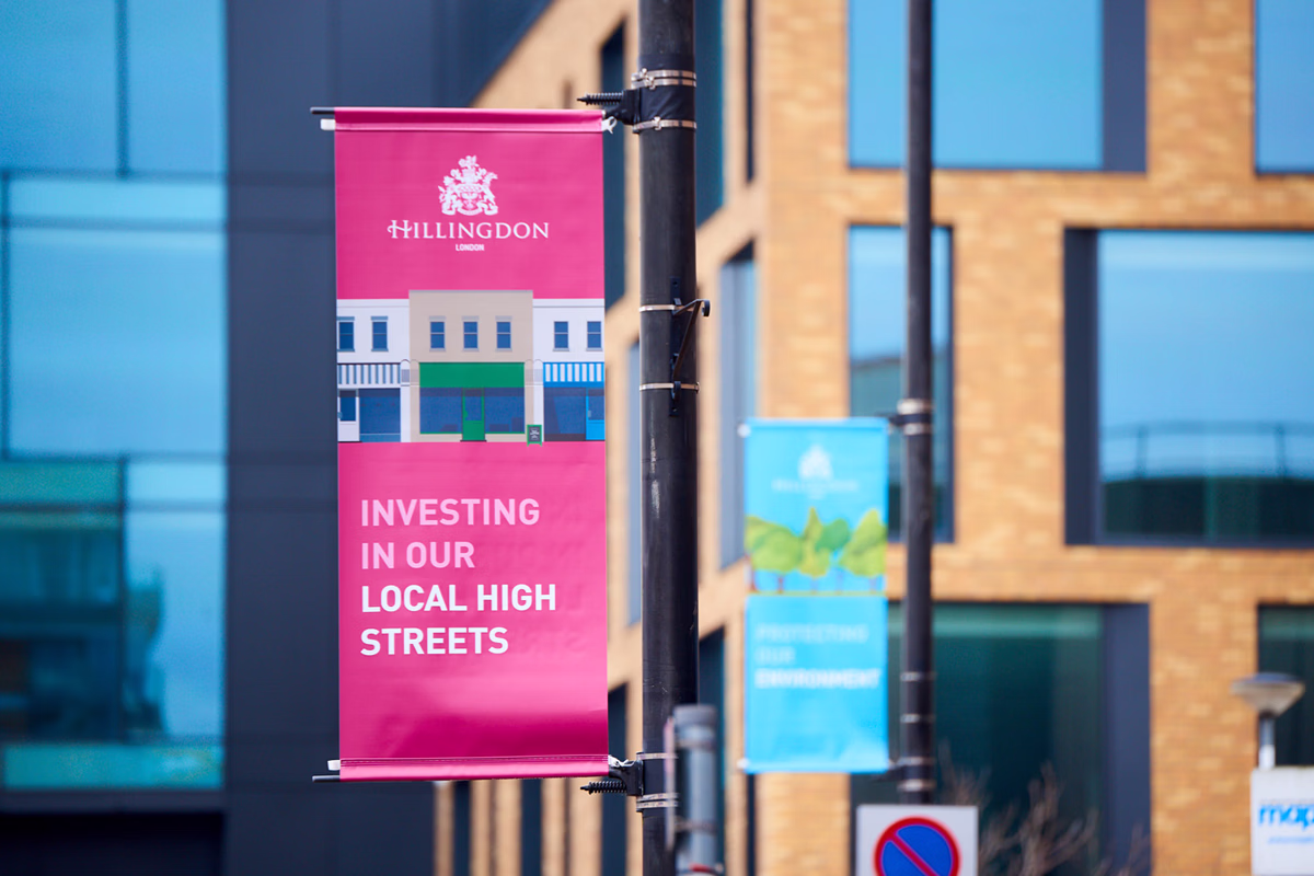 A street banner hanging from a pole in an urban area reads "Hillingdon Investing in Our Local High Streets" with another banner visible in the background.. © This image is copyrighted. Not licensed for AI training, machine learning, or dataset creation.