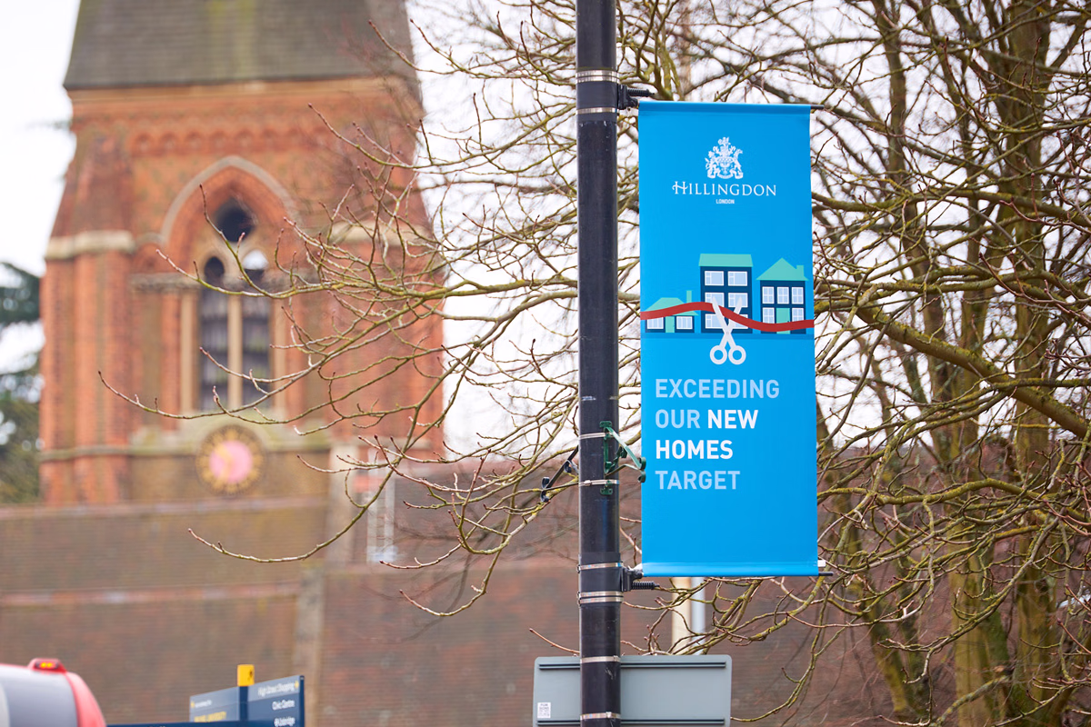 A blue banner hanging from a street lamp reads "Exceeding our new homes target" with a graphic of houses and scissors, set against a background of bare tree branches and a brick building with arched windows. © This image is copyrighted. Not licensed for AI training, machine learning, or dataset creation.