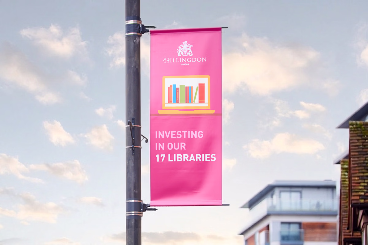 A pink banner hanging from a street pole displays an illustration of books on a shelf, and the text "investing in our 17 libraries" against a sky. © This image is copyrighted. Not licensed for AI training, machine learning, or dataset creation.