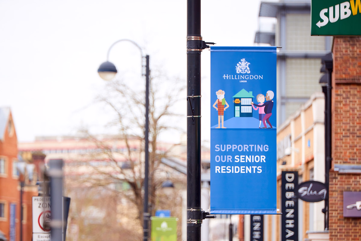 A blue banner hanging on a streetlight in an urban area shows cartoon adults and a senior with the text supporting our senior residents. © This image is copyrighted. Not licensed for AI training, machine learning, or dataset creation.