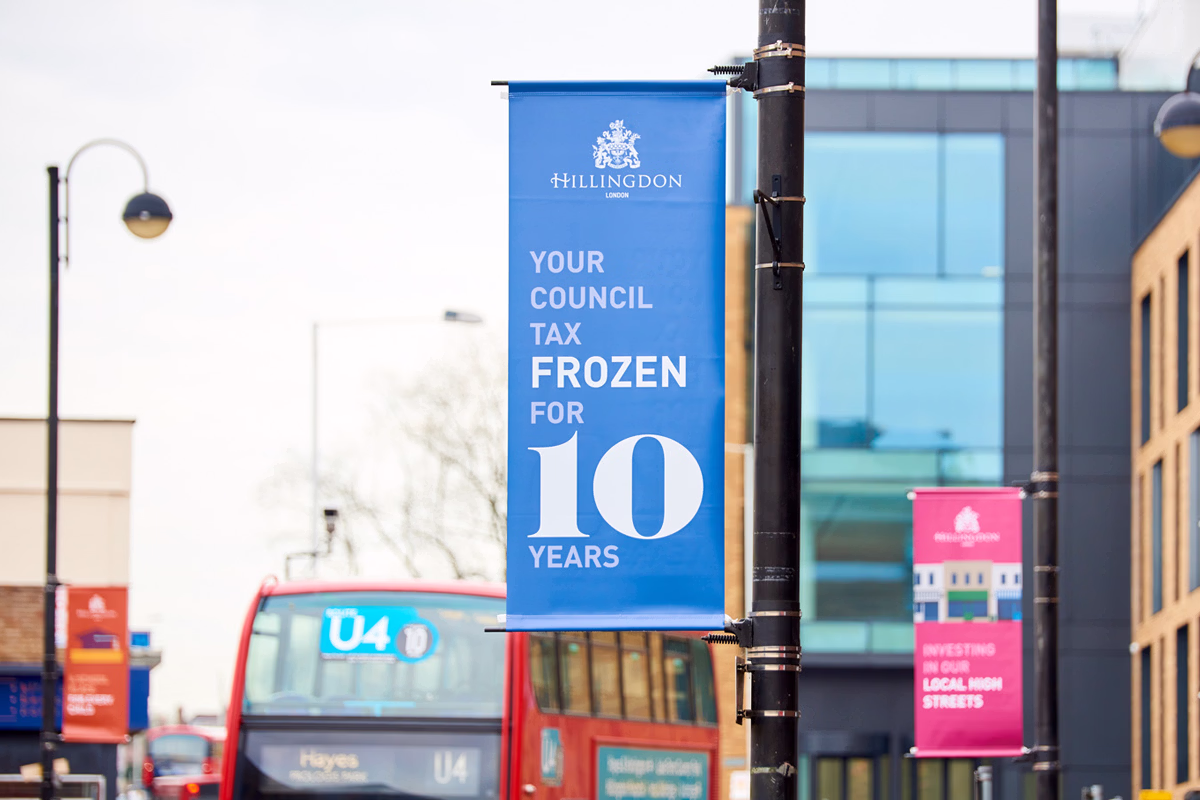 A blue street banner attached to a pole in an urban area reads that the council tax is frozen for ten years, with buses and buildings visible in the background. © This image is copyrighted. Not licensed for AI training, machine learning, or dataset creation.